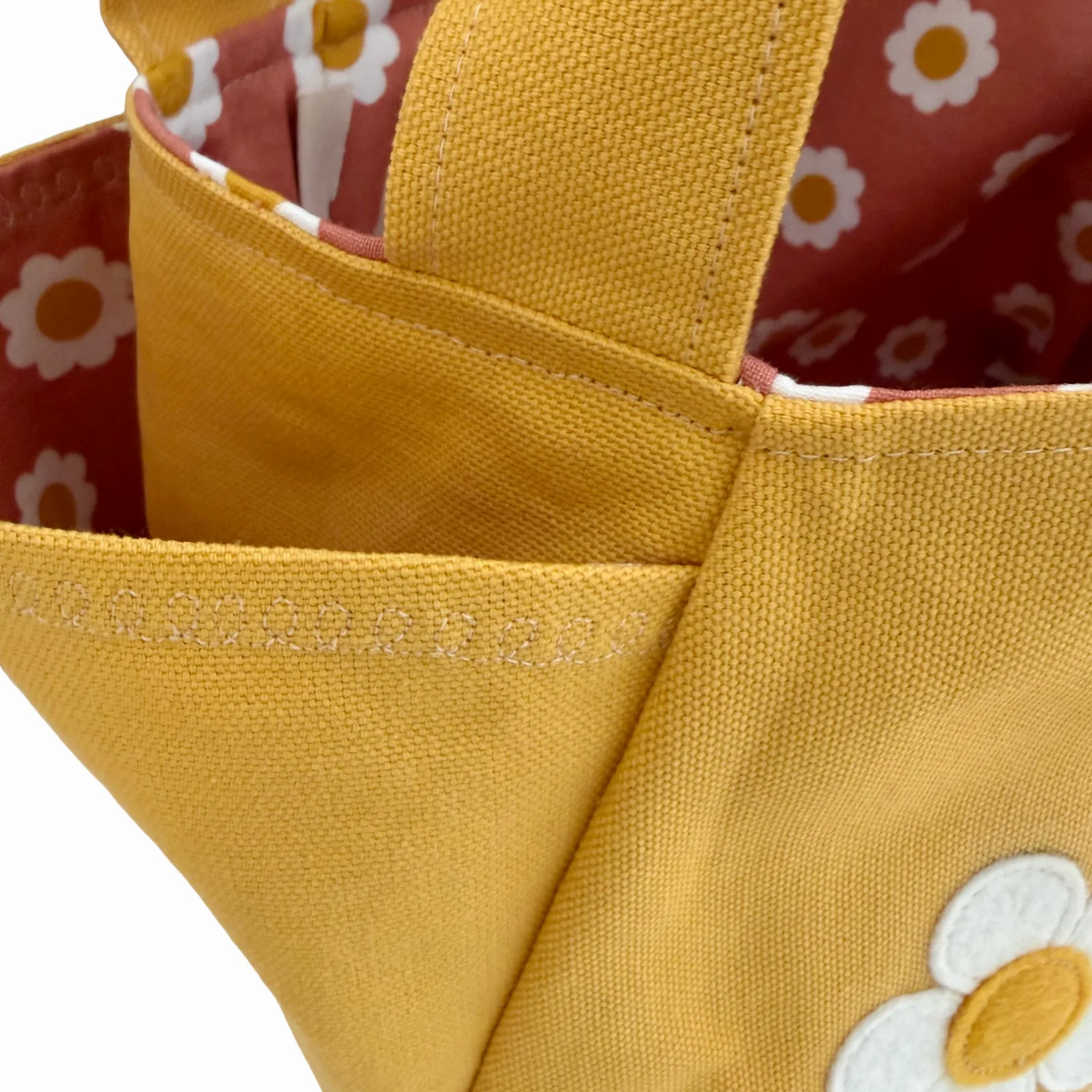Close-up of a mustard yellow canvas tote bag with a daisy applique on the front and a red floral lining visible inside.