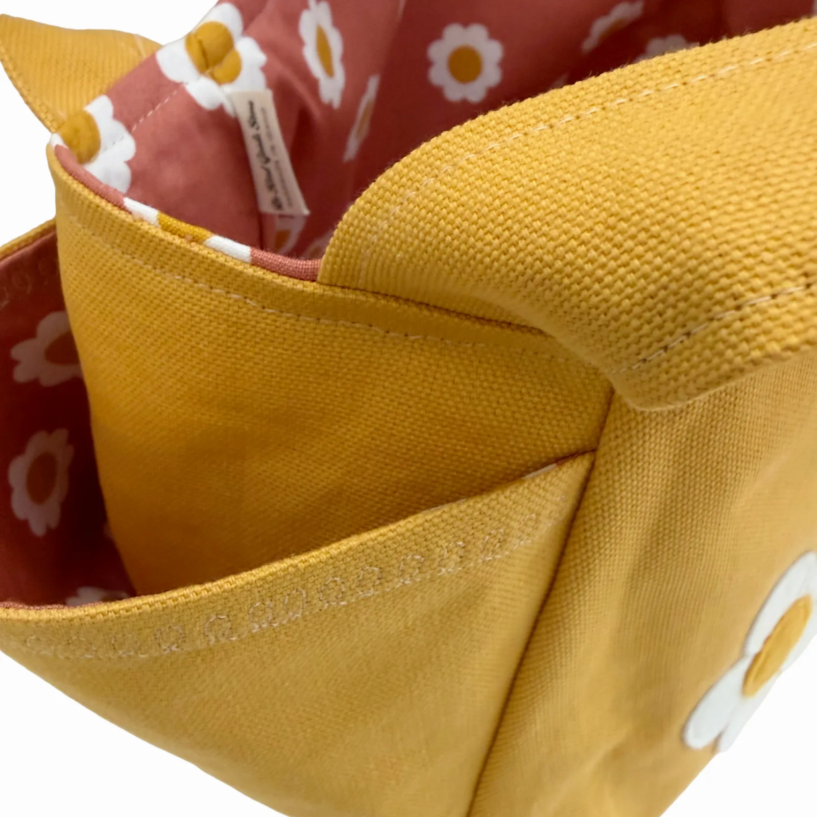 Close-up of a mustard yellow canvas tote bag with a daisy pattern on the exterior and a dusty pink lining with white daisy prints inside.