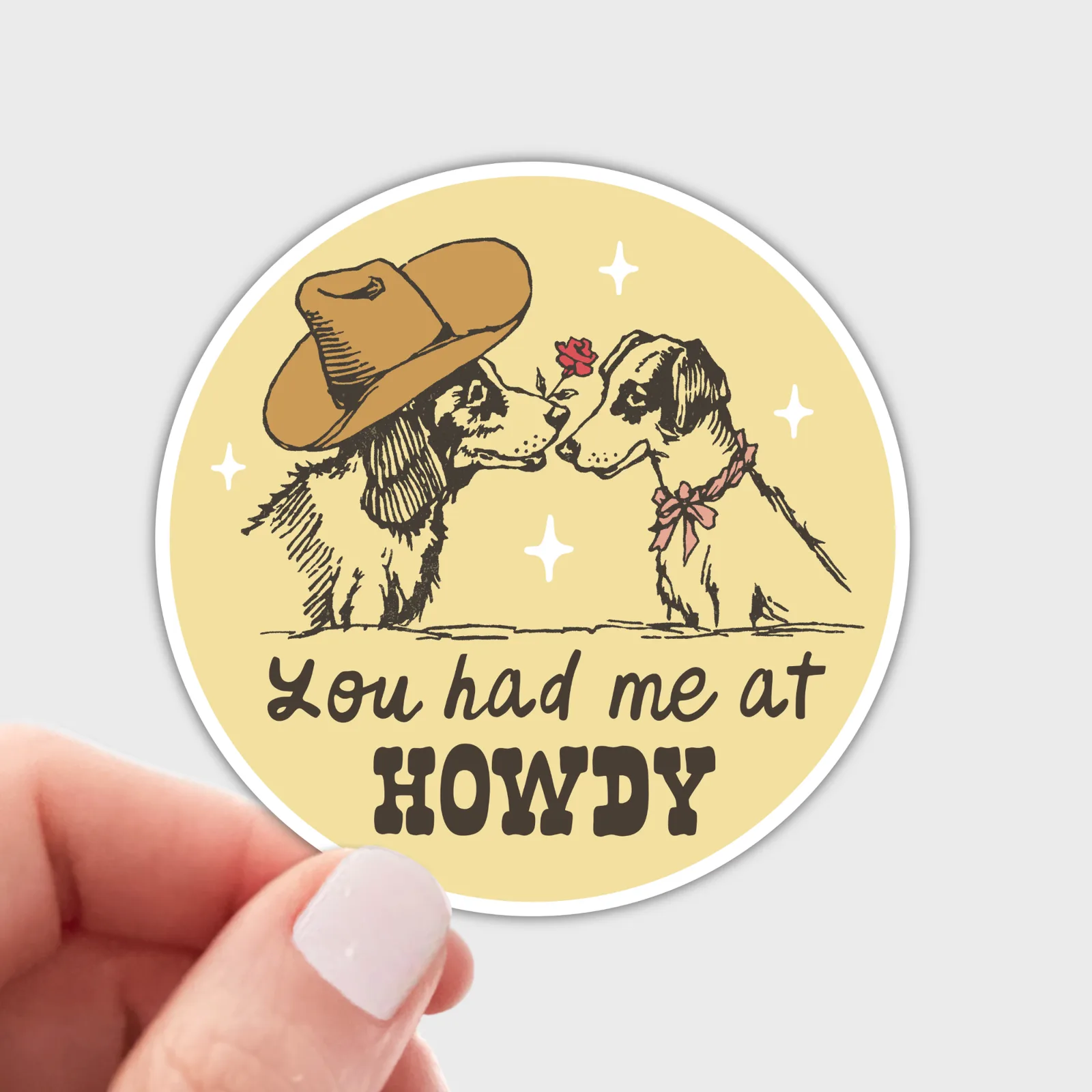 A round sticker held by a hand against a light gray background. The sticker features a vintage-style illustration of two dogs kissing, one wearing a brown cowboy hat and the other wearing a pink bow tie, with the text 'You had me at HOWDY' on a pale yellow background.
